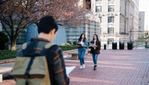 Two female students walking on a college campus, with a male student in the foreground